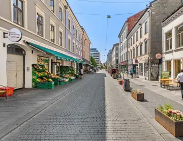 Handlegaten Torggata, sett fra øst til vest. Gågaten er pyntet med blomsterkasser og brostein. En grønnsakshandel på venstre side og restauranter med uteservering på høyre side.
