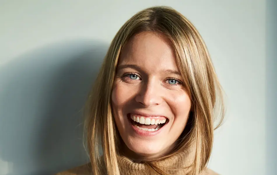 En dame med brun strikkegenser står foran en hvit vegg og smiler