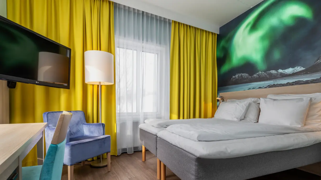 Dobbeltrom med twin senger, lilla lenestol, skrivebord med turkis stol, gule gardiner og stort nordlys bilde på veggen.