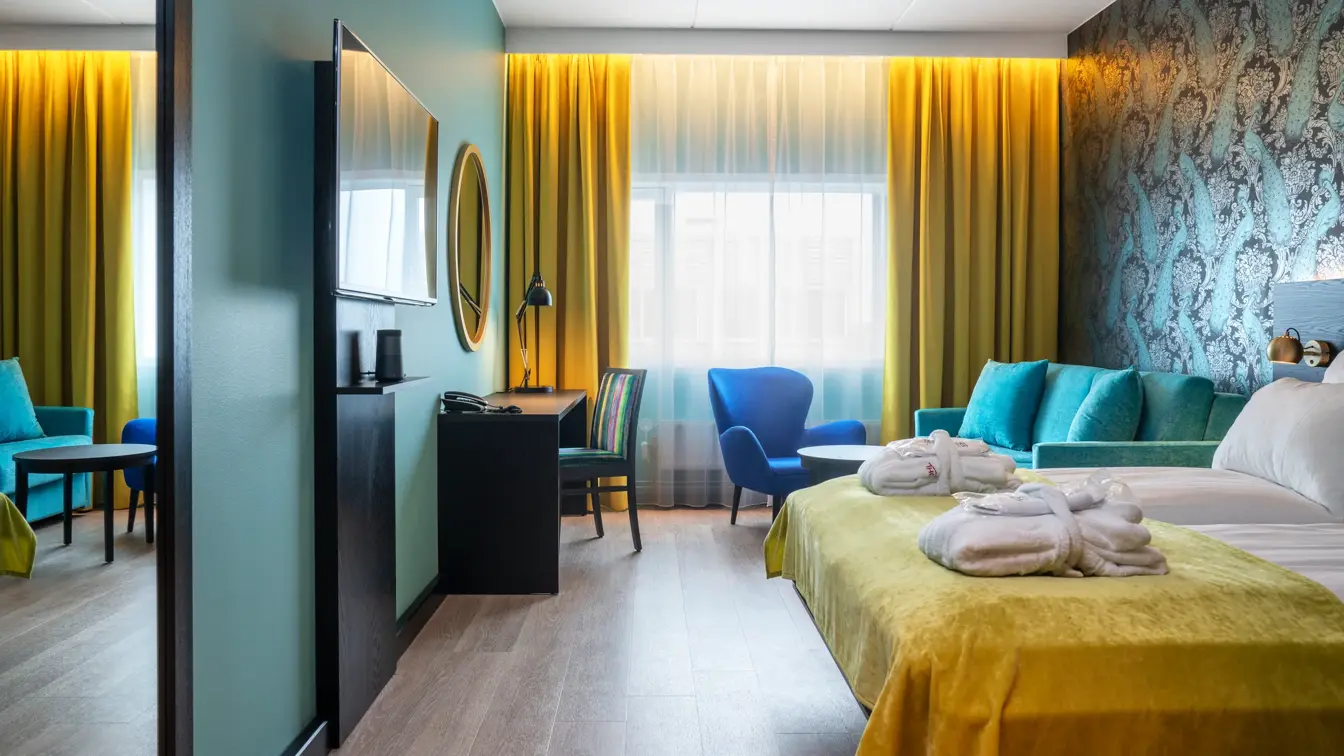 Vindu fra gulv til tak med gule gardiner, turkis vegg, tapetvegg med mønster, turkis sofa, blå stol, dobbeltseng, TV og speil på vegg. 
