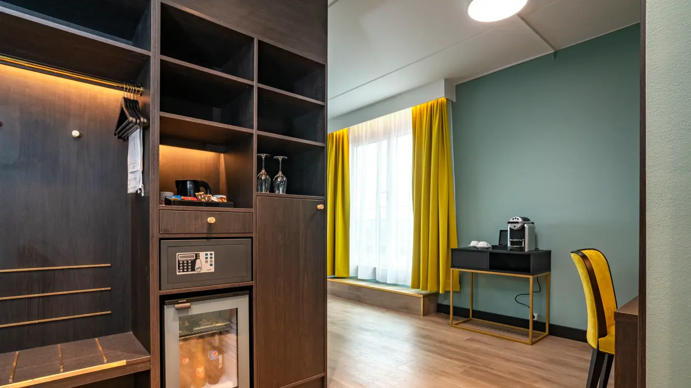 Skap, skrivebord, gul lenestol, vindu, grønne/turkise vegger, kaffemaskin, minibar