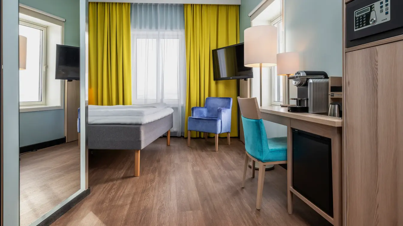 Dobbeltrom med dobbeltseng, gule gardiner, lilla lenestol, skrivebord med turkis stol, Nespresso maskin og safe.