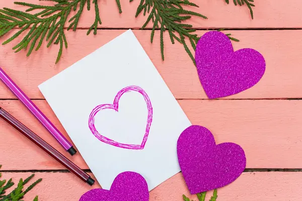 Hvitt kort med tegnede, lilla hjerter ligger på et rosa bord. Foto tatt ovenfra.
