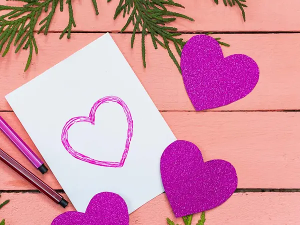 Hvitt kort med tegnede, lilla hjerter ligger på et rosa bord. Foto tatt ovenfra.