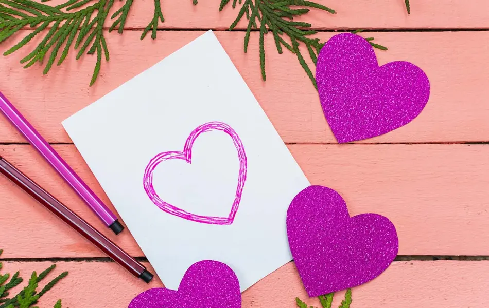Hvitt kort med tegnede, lilla hjerter ligger på et rosa bord. Foto tatt ovenfra.