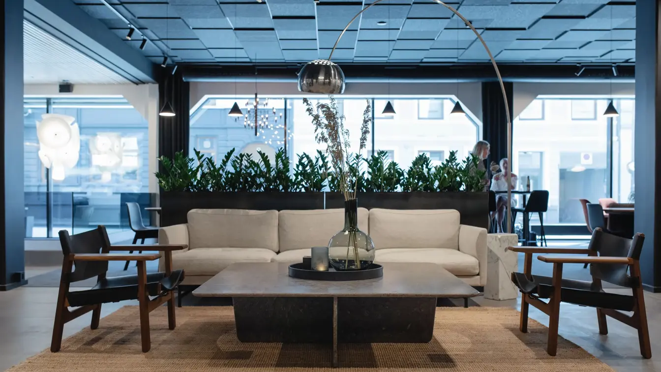 Hvit sofa og mørke stoler rundt et lavt sort bord. Store vinduer ut mot gata og grønne vekster. I lobbyen på Thon Hotel Norge i Kristiansand.