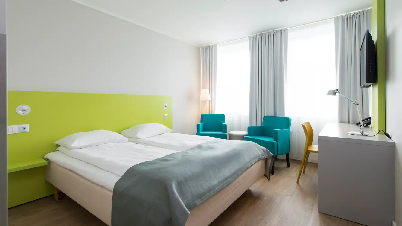 Standard Room Double på Thon Hotel Trondheim. Dobbeltseng med grått sengeteppe. Limegrønn sengegavl. Grått skrivebord med tilhørende gul stol. To turkise lenestoler og et rundt bord. 