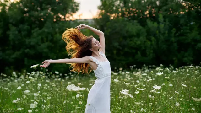 En kvinne med brunt hår står i en eng med blomster med armene ut og i en hvit kjole.