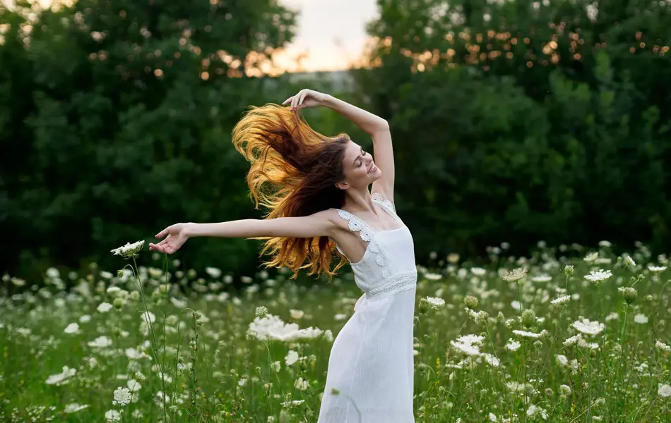 En kvinne med brunt hår står i en eng med blomster med armene ut og i en hvit kjole.