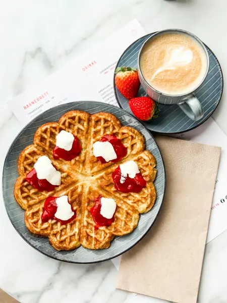 Tallerken med vaffel med syltetøy og rømme og en kaffe latte på et bord.