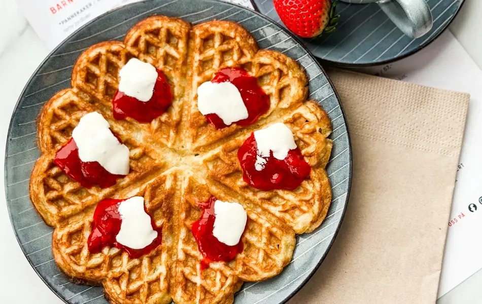 Tallerken med vaffel med syltetøy og rømme og en kaffe latte på et bord.