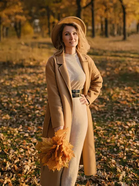 Dame med brun kjole og brun kåpe med mørkebrunt belte i midjen poserer med gule blader utendørs