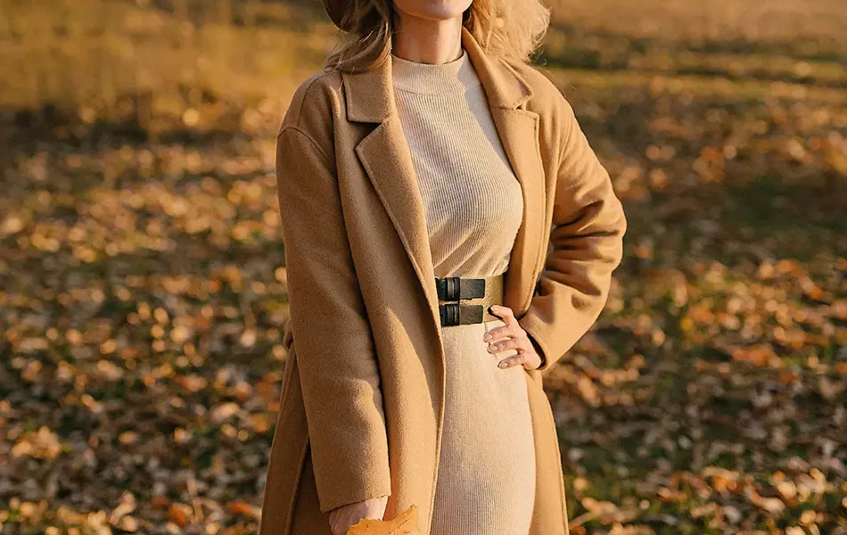 Dame med brun kjole og brun kåpe med mørkebrunt belte i midjen poserer med gule blader utendørs