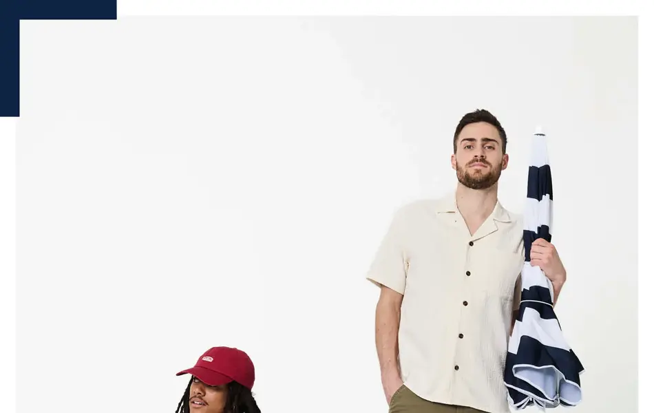 To unge menn i t-skjorte og shorts, en sitter i en stol og en står med en parasoll.