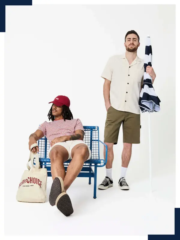 To unge menn i t-skjorte og shorts, en sitter i en stol og en står med en parasoll.