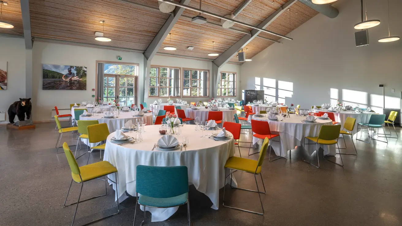 Selskapslokale dekket opp med runde bord, hvite duker, fargerike stoler, vinduer og høyt under taket.