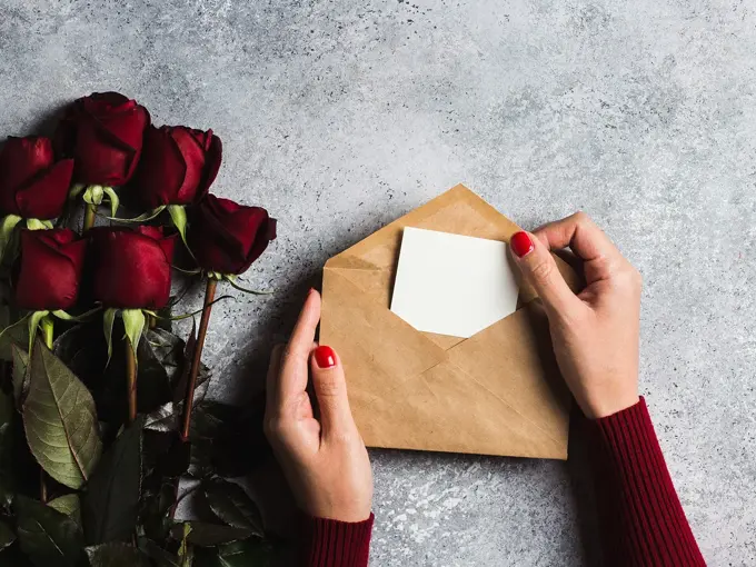 Valentines day women hand holding envelope with greeting card mothers day red rose gift surprise on grey background with copyspace. Love flower gift for woman on romantic holiday birthday wedding