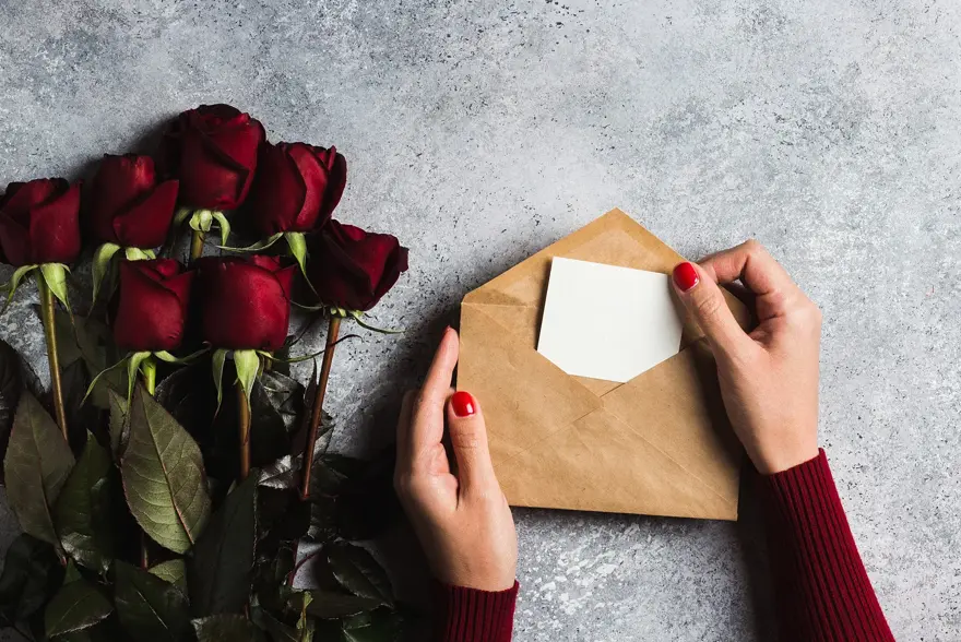Valentines day women hand holding envelope with greeting card mothers day red rose gift surprise on grey background with copyspace. Love flower gift for woman on romantic holiday birthday wedding