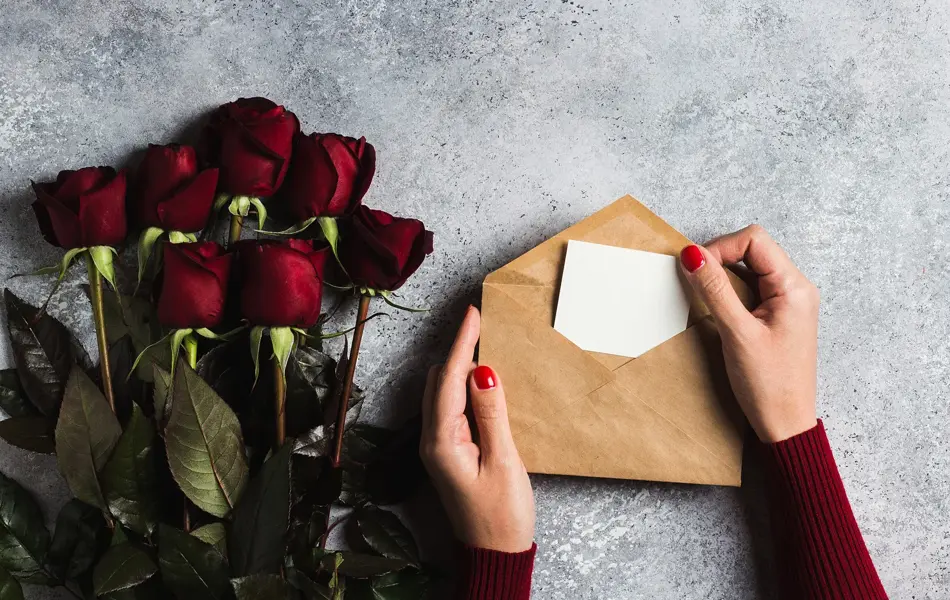 Valentines day women hand holding envelope with greeting card mothers day red rose gift surprise on grey background with copyspace. Love flower gift for woman on romantic holiday birthday wedding