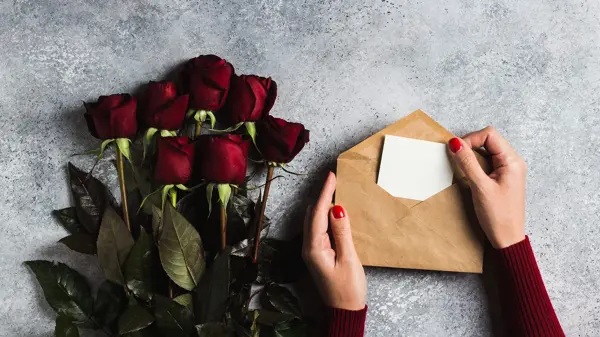 Valentines day women hand holding envelope with greeting card mothers day red rose gift surprise on grey background with copyspace. Love flower gift for woman on romantic holiday birthday wedding