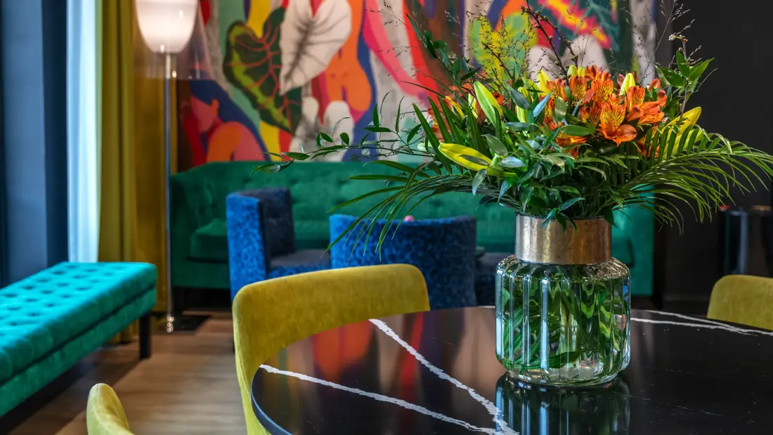 Nærbilde av en fargerik blomsterbukett i vase på et rundt bord med gule stoler rundt. I bakgrunnen er det fargerike møbler og en vegg med fargerik tapet.