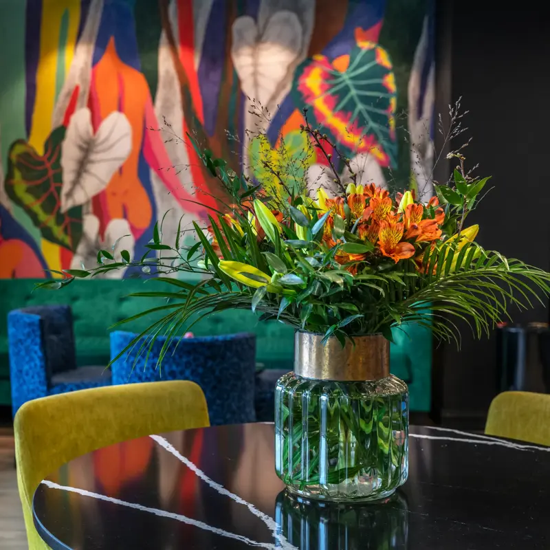 Nærbilde av en fargerik blomsterbukett i vase på et rundt bord med gule stoler rundt. I bakgrunnen er det fargerike møbler og en vegg med fargerik tapet.