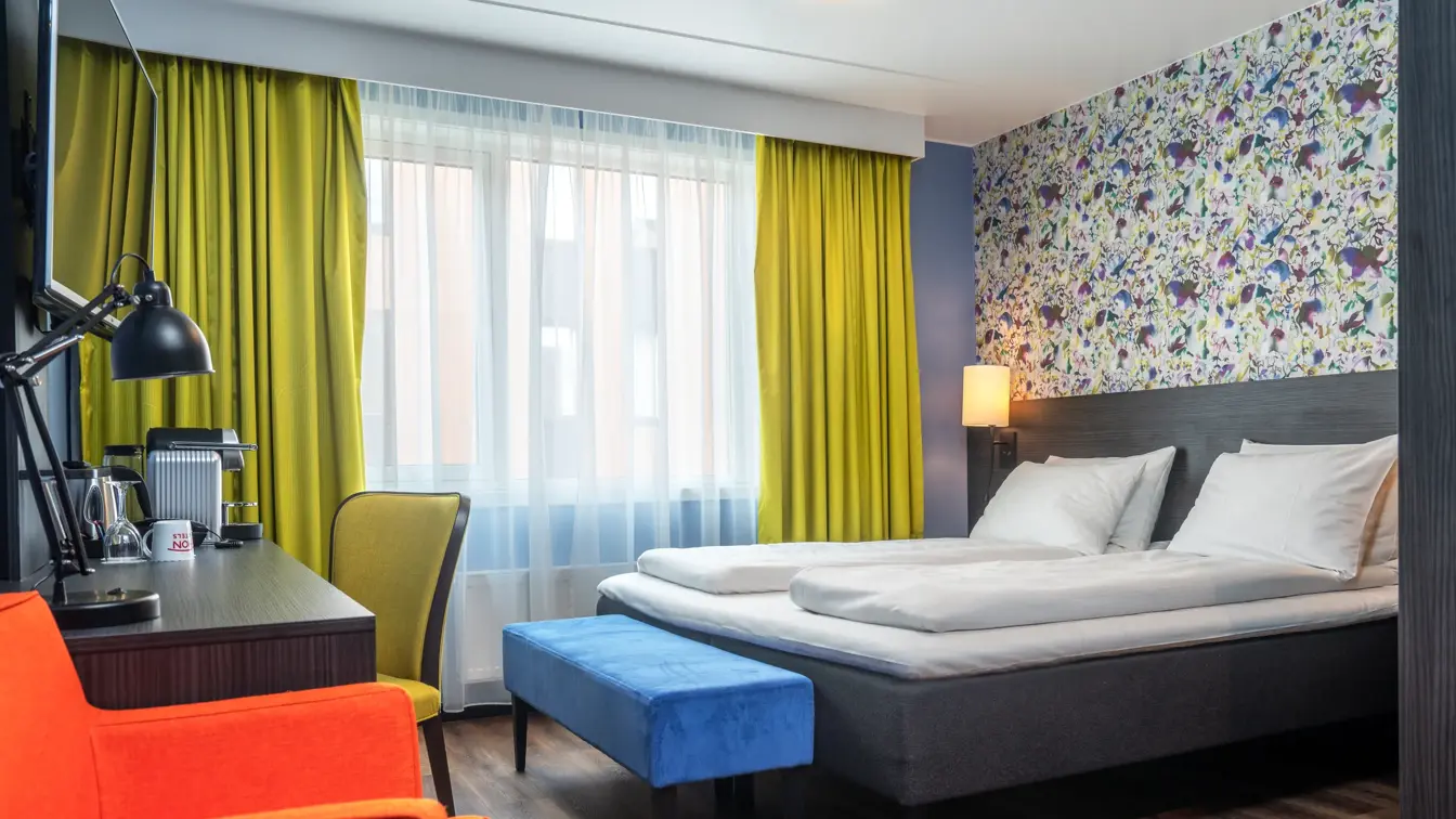 Gule gardiner, dobbeltseng, oransj stol, skrivepult, vindu, blomstertapet, gul kontorstol, parkettgulv. 