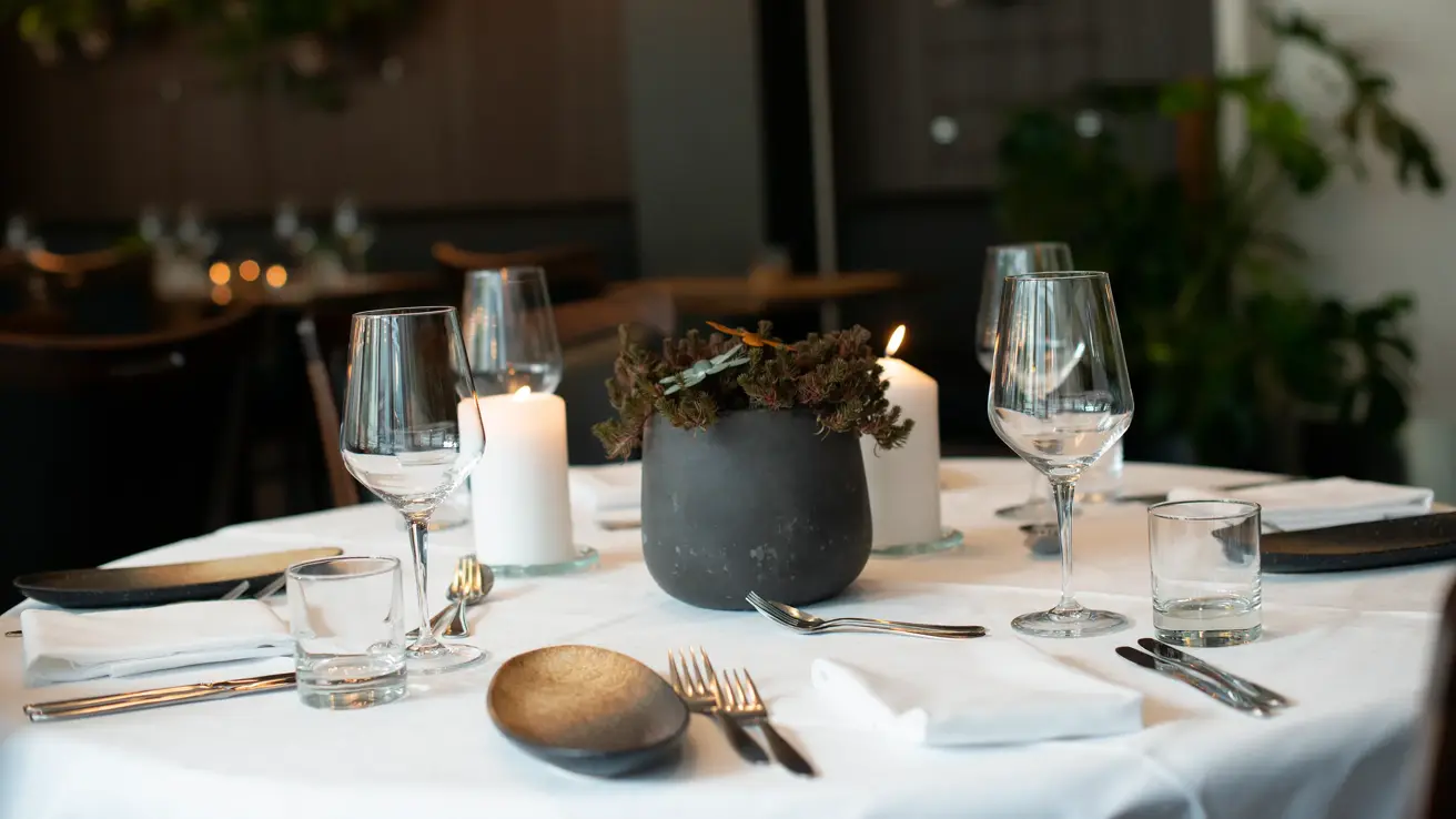 Bord dekket med hvit duk og servise. Svart blomsterpotte stående på midten av bordet. 