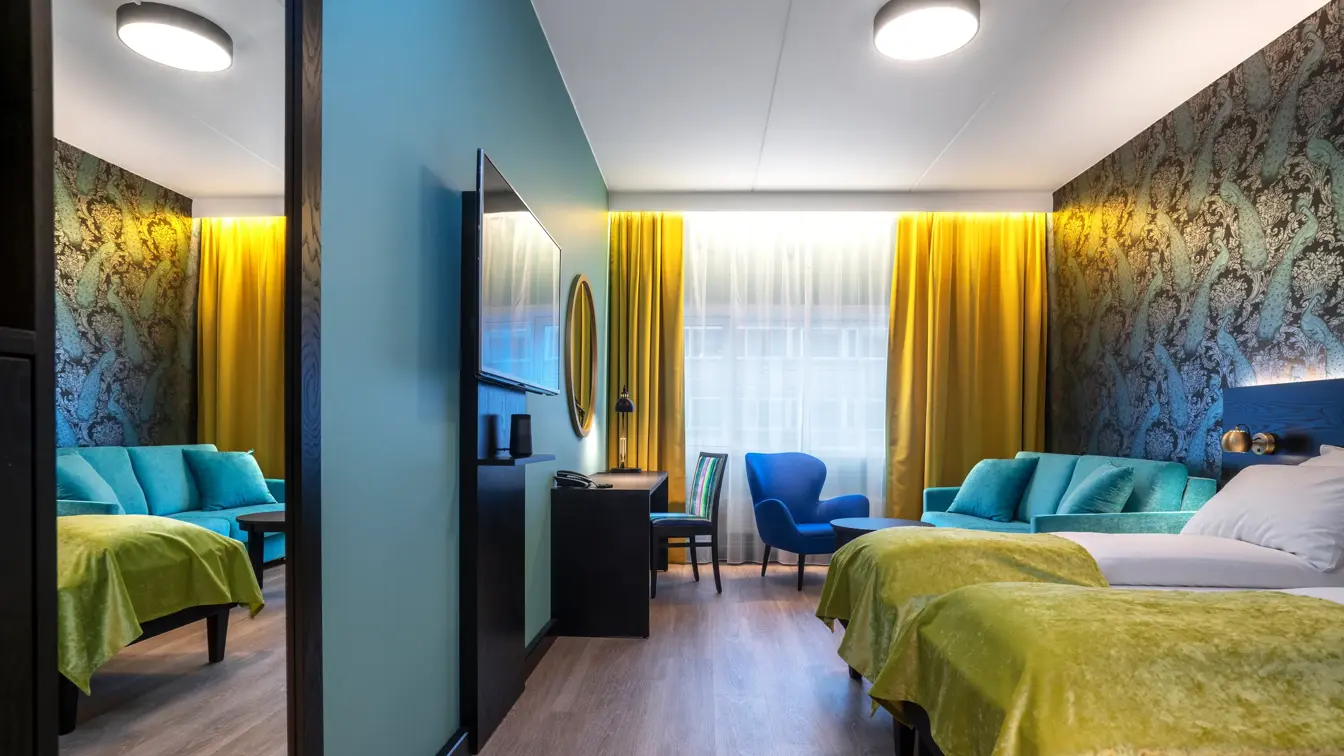 Parkettgulv, vindu fra gulv til tak, gule gardiner, veggspeil, to enkle senger, tapetvegg med mønster, skrivebord, to stoler, TV, turkis sofa. 