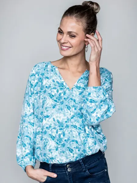 Smilende dame med lyst hår, satt opp i en bun. Har på seg turkisblomstret bluse og mørkeblåe jeans. 