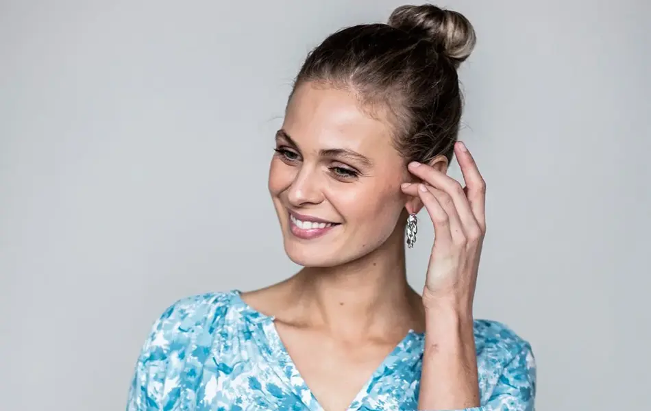 Smilende dame med lyst hår, satt opp i en bun. Har på seg turkisblomstret bluse og mørkeblåe jeans. 