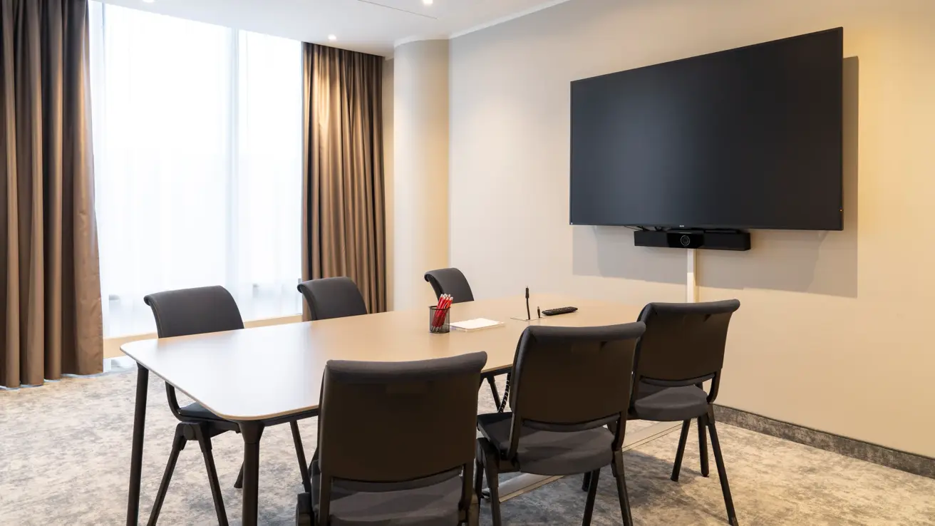 Lyst møterom med langbord og seks stoler rundt, TV skjerm på veggen, stort vindu med gardiner.