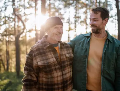 To smilende menn med rutete og grønn jakke går i skogen med trær og lysning i bakgrunnen 