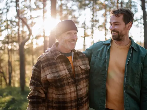 To smilende menn med rutete og grønn jakke går i skogen med trær og lysning i bakgrunnen 