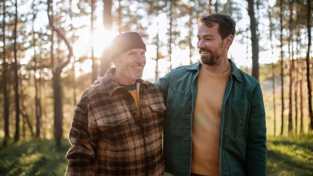 To smilende menn med rutete og grønn jakke går i skogen med trær og lysning i bakgrunnen 