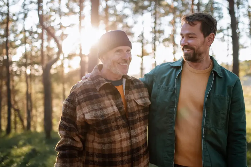 To smilende menn med rutete og grønn jakke går i skogen med trær og lysning i bakgrunnen 