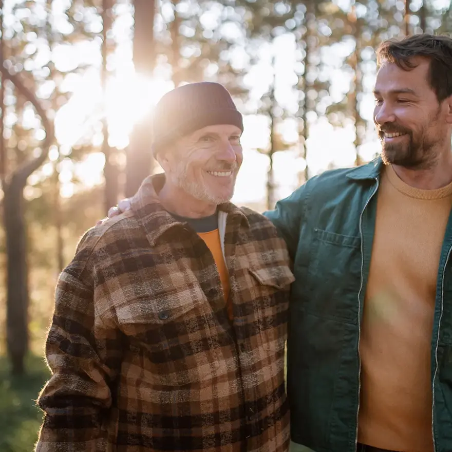 To smilende menn med rutete og grønn jakke går i skogen med trær og lysning i bakgrunnen 