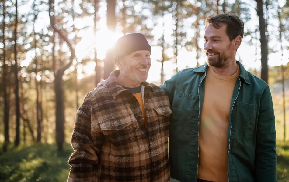To smilende menn med rutete og grønn jakke går i skogen med trær og lysning i bakgrunnen 
