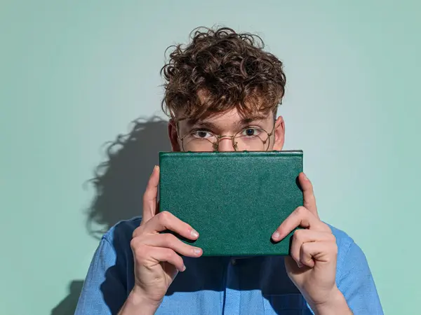 Gutt med briller og blå skjorte holder grønn bok foran nedre del av ansiktet sitt