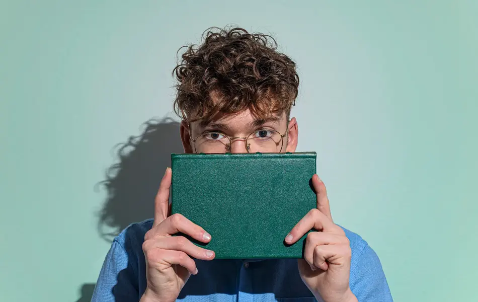 Gutt med briller og blå skjorte holder grønn bok foran nedre del av ansiktet sitt