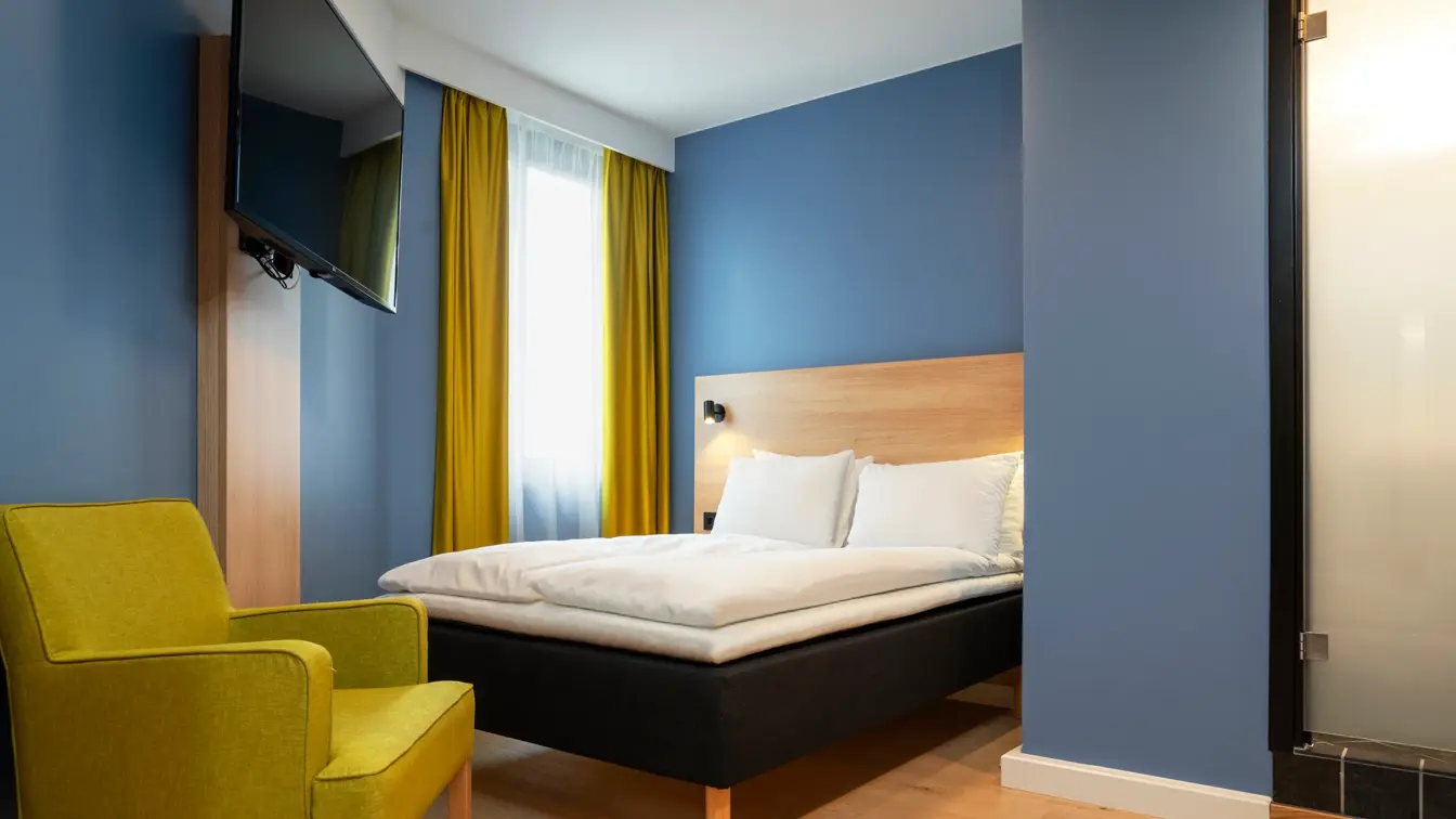 Gul stol, blå vegger, TV og queen seng på standard rom queen på Thon Hotel Astoria.