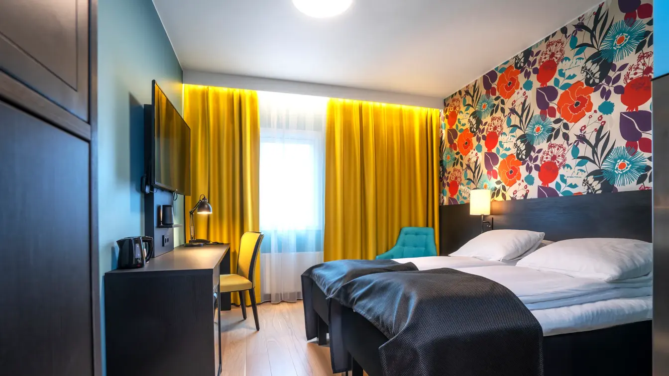 Blomstrete tapet, et stort vindu fra gulv til tak, gule gardiner, parkettgulv, to enkle senger, svart skrivebord, en gul stol og en blå stol. 