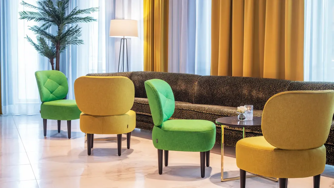 Resepsjon, lobby, store vinduer fra gulv til tak, en mørk lang sofa plassert inntil vinduene, fire stoler i fargen grunn og gul.