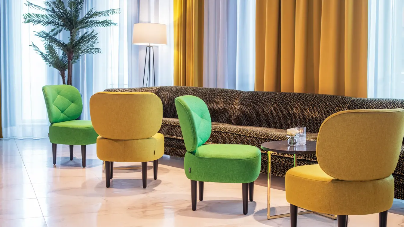 Resepsjon, lobby, store vinduer fra gulv til tak, en mørk lang sofa plassert inntil vinduene, fire stoler i fargen grunn og gul.