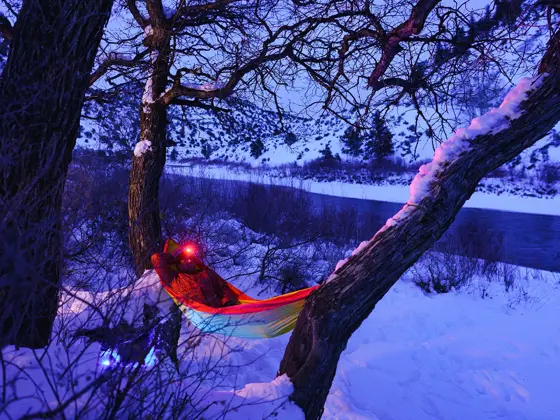 Man Relaxing at Campsite Swinging in Hammock at Dusk - Recreation in winter with man enjoying a peaceful tranquil setting along majestic river in hammock wearing headlamp at dusk during blue hour. Outdoor adventure series.