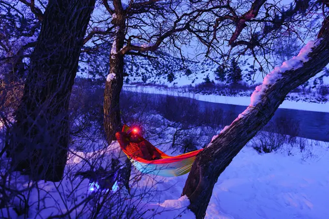 Man Relaxing at Campsite Swinging in Hammock at Dusk - Recreation in winter with man enjoying a peaceful tranquil setting along majestic river in hammock wearing headlamp at dusk during blue hour. Outdoor adventure series.
