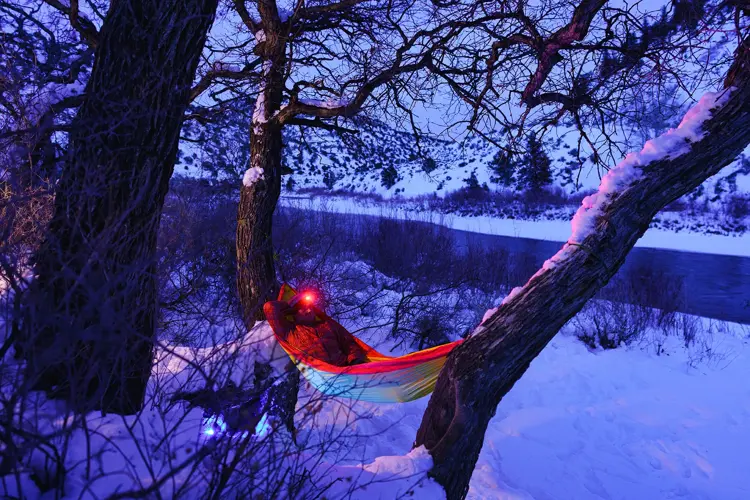 Man Relaxing at Campsite Swinging in Hammock at Dusk - Recreation in winter with man enjoying a peaceful tranquil setting along majestic river in hammock wearing headlamp at dusk during blue hour. Outdoor adventure series.