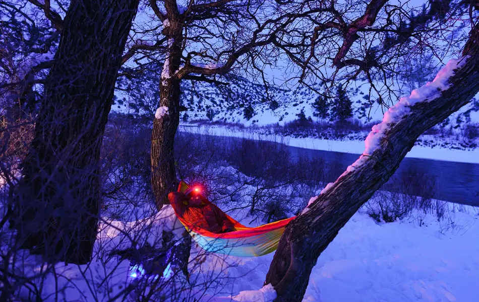 Man Relaxing at Campsite Swinging in Hammock at Dusk - Recreation in winter with man enjoying a peaceful tranquil setting along majestic river in hammock wearing headlamp at dusk during blue hour. Outdoor adventure series.