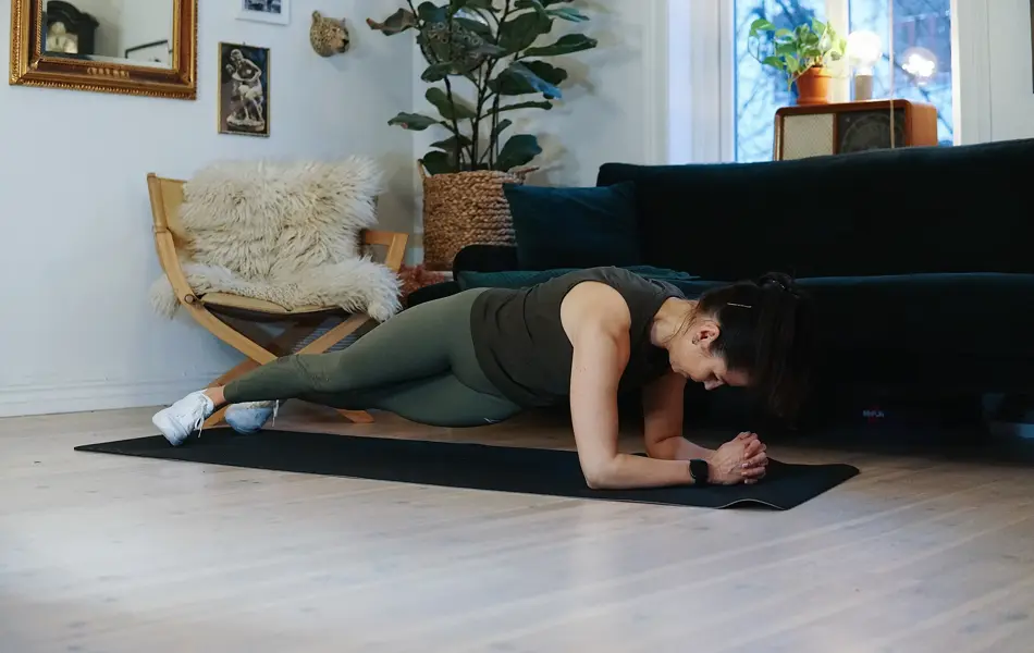 treningsekspert Anna Johansen trener på matte på gulvet, mørkegrønn sofa i bakgrunnen, lys stol med pels oppi, str grønn plante, bilder på veggen