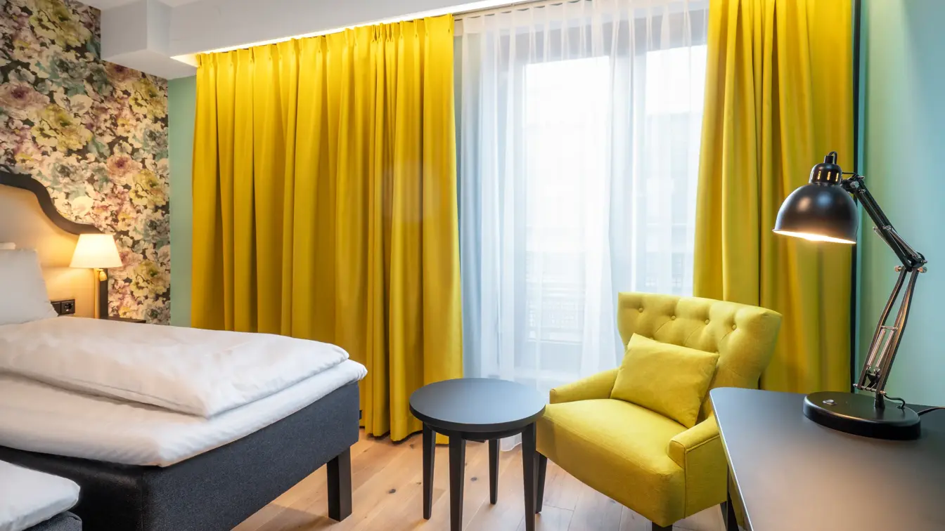 To enkeltsenger, blomstret tapet bak sengene, gul stol, arbeidspult og store vinduer med store gule gardiner på twin room på Thon Hotel Cecil i Oslo sentrum